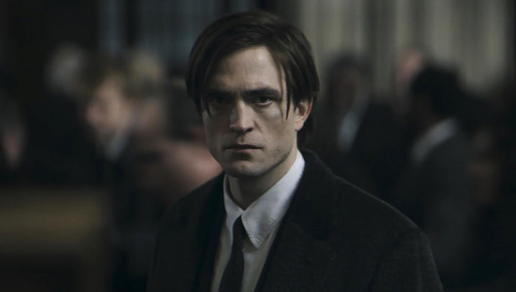 Bruce Wayne, wearing a black suit and white dress shirt, stands with his back to a crowd of people. He is in sharp focus, while the crowd blurs together, and he has a grim expression on his face as he gazes at something off-screen.