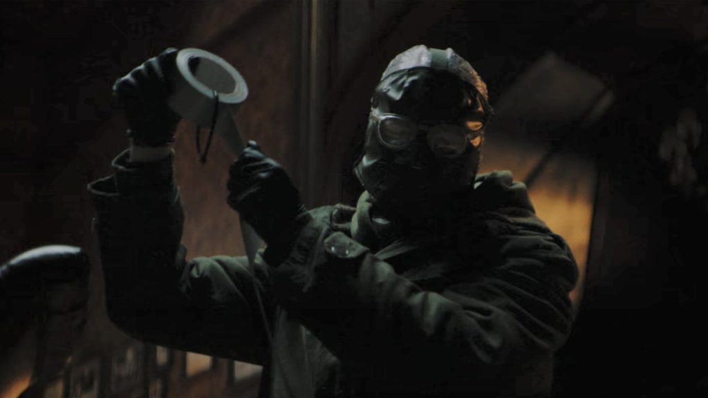 The Riddler, head covered by what appears to be a leather mask with glasses, holds a roll of silver duct tape, in the process of ripping off a strip.