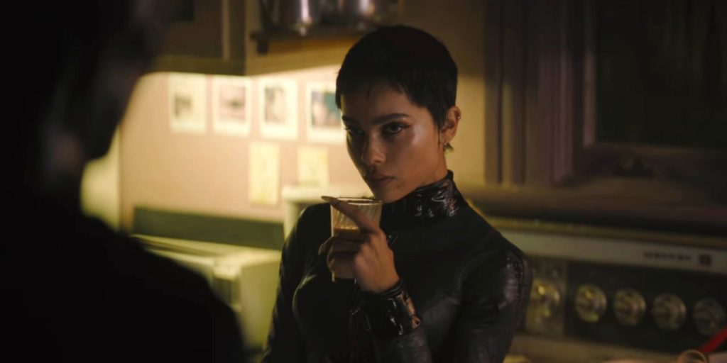 Selina Kyle/Catwoman leans against her kitchen counter, glass of milk in one hand. She faces Batman with a serious expression.